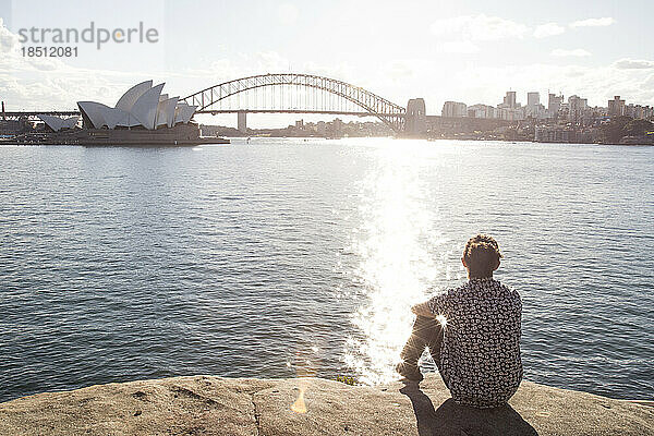 Mann mit Dutt sitzt am Rande des Wassers und starrt auf den Hafen von Sydney