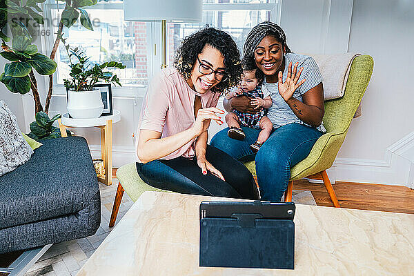 Lächelnde Lesben mit Baby genießen zu Hause Videoanrufe auf einem digitalen Tablet