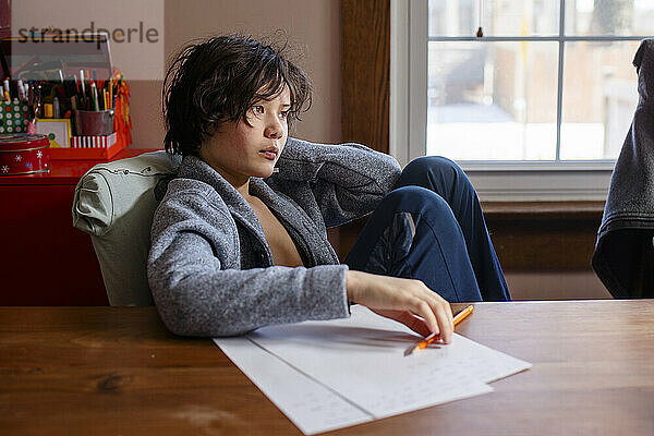 Ein Junge in Strickjacke sitzt am Tisch am Fenster und macht Hausaufgaben