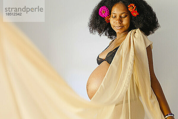 Eine schwangere Frau mit Blumen im Haar steht in goldenem Stoff gehüllt