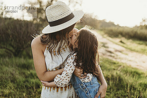 Junge Mutter mit Hut und Tochter küssen sich auf einer Wiese im Gegenlicht