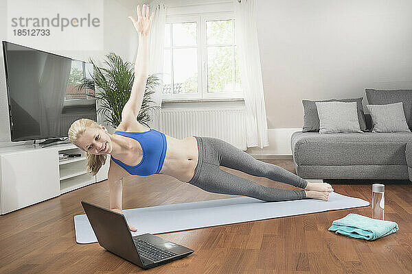 Junge Frau mit Laptop macht Sideplank-Pose auf einer Trainingsmatte im Wohnzimmer  Bayern  Deutschland