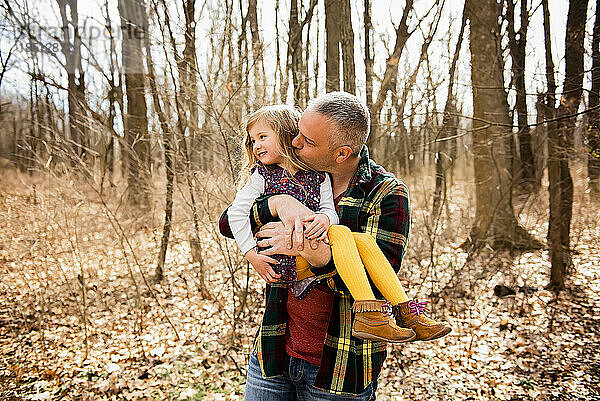 Liebevoller Vater hält und küsst lächelnde Tochter im Herbstwald auf die Wange