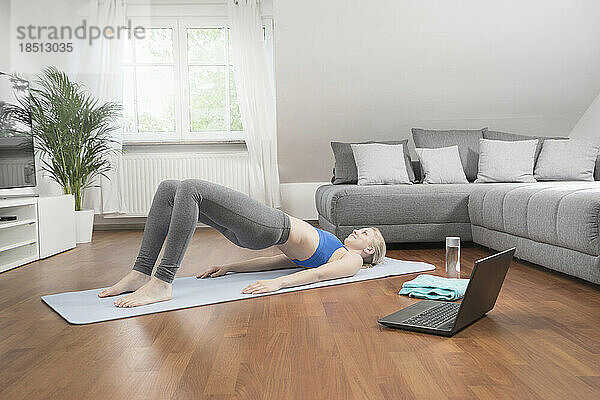 Junge Frau mit Laptop macht Yoga auf einer Gymnastikmatte im Wohnzimmer  Bayern  Deutschland