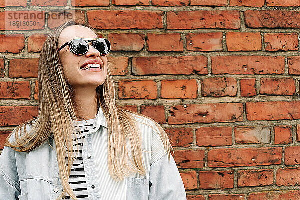 Langhaarige Frau mit Sonnenbrille lacht in der Nähe einer Ziegelmauer