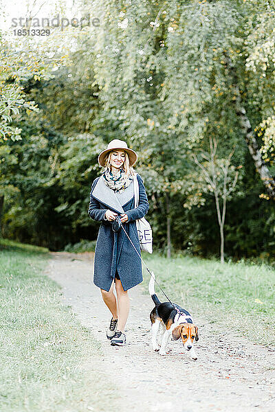 Junge Frau in Mantel und Hut geht mit einem Beagle-Hund im Park spazieren