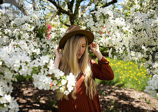 Junge Frau schaut nach unten und hält einen Hut  umgeben von weißen Birnenblumen