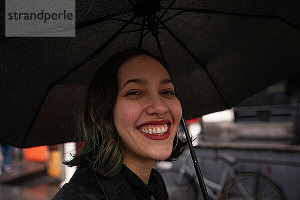 Lächelnde junge Frau unter Regenschirm  die an einem regnerischen Tag spaziert.