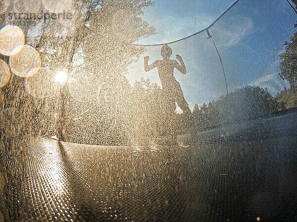 Silhouette eines Jungen auf einem Trampolin mit Wasserspritzern und Blendenfleck