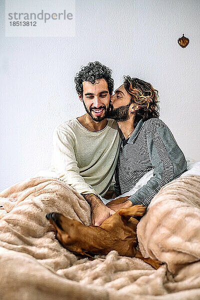 Schwuler Mann küsst Freund  der zu Hause mit Hund im Bett spielt