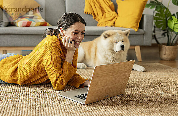 Lächelnde Frau  die zu Hause auf dem Teppich liegt und den Laptop neben dem Hund benutzt