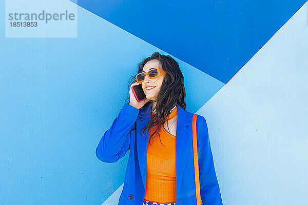 Fröhliche  modische Frau mit Sonnenbrille  die vor einer blauen Wand mit ihrem Smartphone spricht