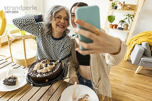 Glückliche reife Frau hält Kuchen und Tochter macht zu Hause ein Selfie per Smartphone