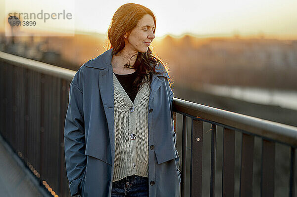 Smiling woman standing on bridge by railing at sunset