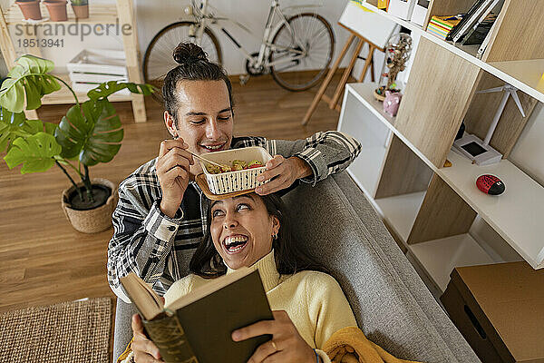 Smiling man eating snack with girlfriend reading book lying on sofa at home
