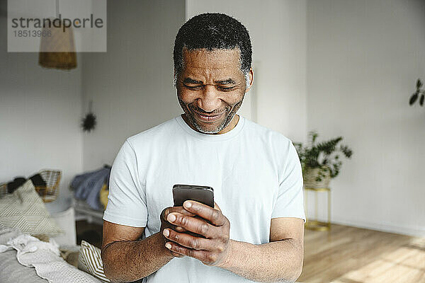 Lächelnder reifer Mann benutzt Mobiltelefon im Wohnzimmer zu Hause