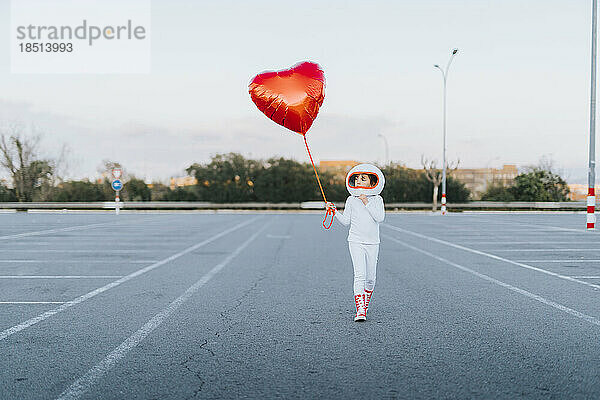 Mädchen im Astronautenkleid geht mit rotem Herzballon auf Parkplatz