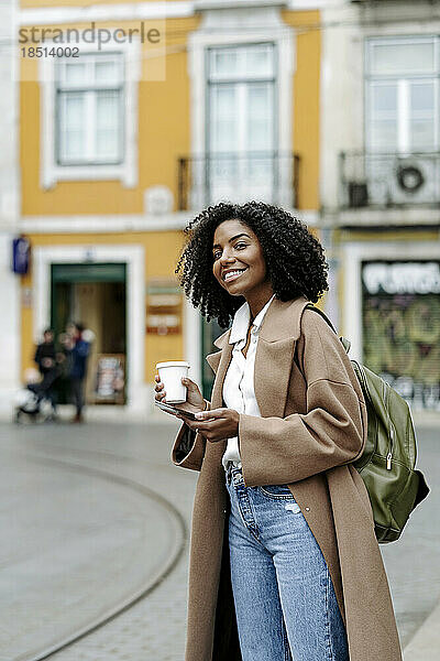 Glückliche junge Frau steht mit Handy und Kaffeetasse an der Straßenbahn
