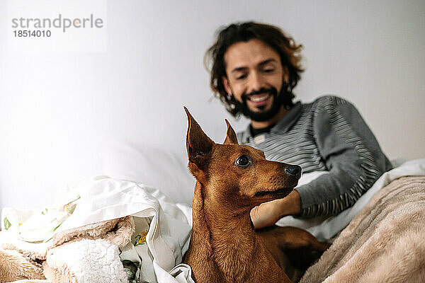 Lächelnder Mann  der zu Hause mit seinem Hund im Bett spielt