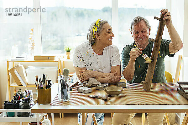 Lächelnde ältere Frau schaut einem Mann zu  der in der Werkstatt Holz poliert