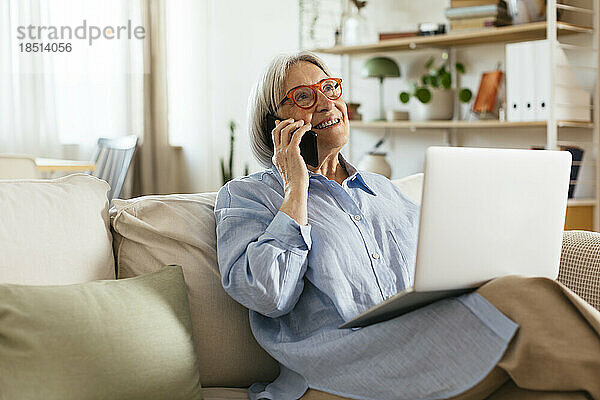 Lächelnde Frau  die mit einem Laptop im Wohnzimmer sitzt und auf einem Smartphone spricht