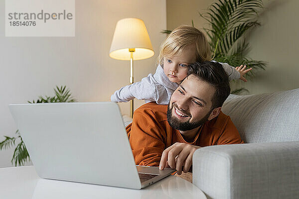 Glücklicher Vater benutzt Laptop  während sein Sohn zu Hause auf dem Vater liegt