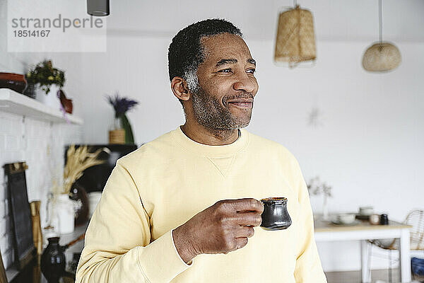 Lächelnder reifer Mann steht mit einer Kaffeetasse zu Hause