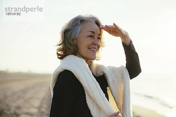 Happy mature woman shielding eyes standing at beach