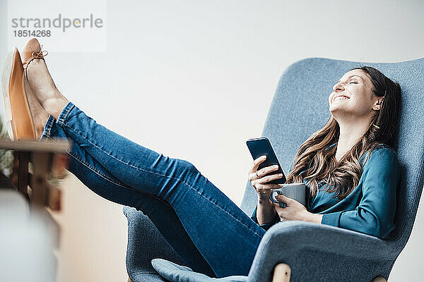 Glückliche junge Frau mit Smartphone entspannt sich zu Hause im Sessel und legt die Füße hoch