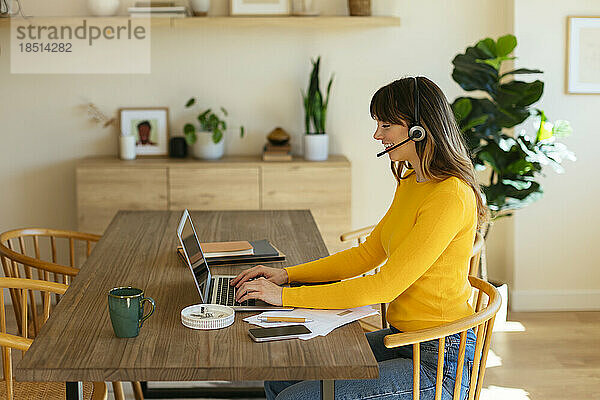 Lächelnder Freiberufler mit Headset  der im Heimbüro am Laptop arbeitet