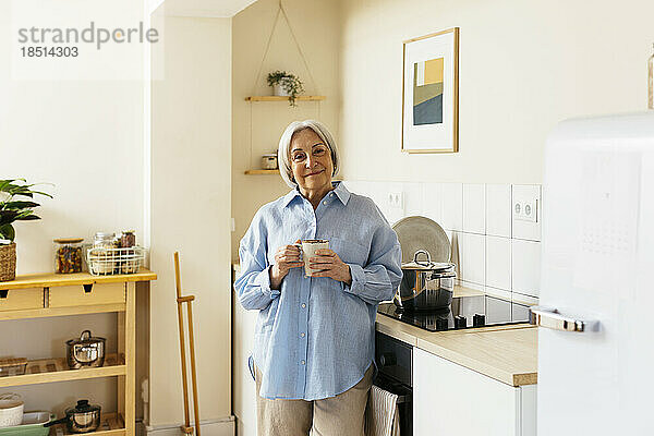 Lächelnde ältere Frau steht mit einer Tasse in der Küche