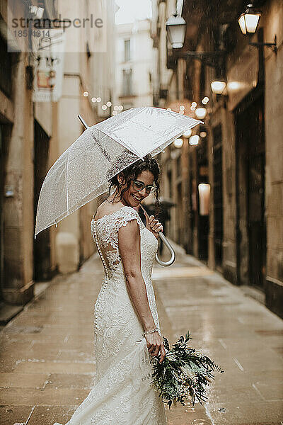 Lächelnde Frau im Hochzeitskleid mit Regenschirm inmitten von Gebäuden