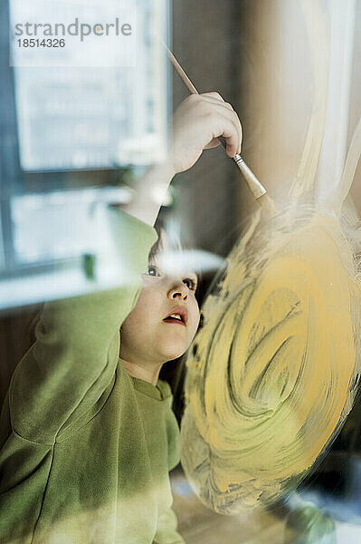 Cute boy drawing sun on glass with paintbrush