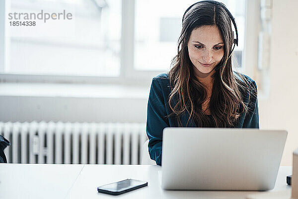 Junge Geschäftsfrau trägt Kopfhörer und arbeitet im Heimbüro am Laptop