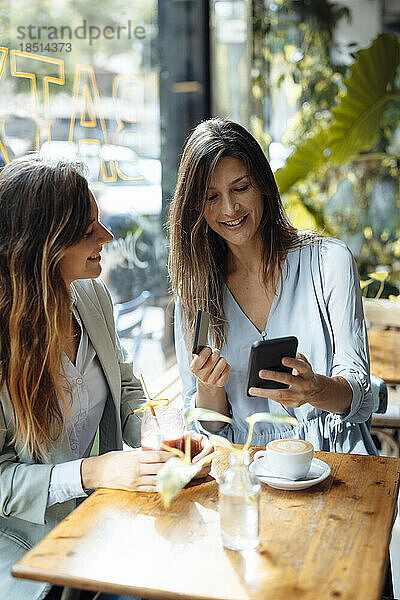 Frau mit Freundin hält Kreditkarte in der Hand und benutzt Smartphone im Café