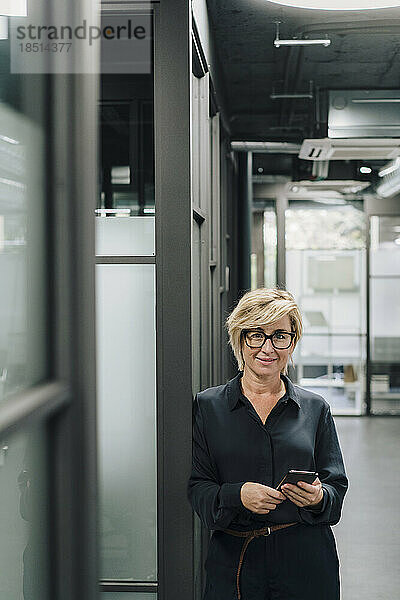 Lächelnde Geschäftsfrau mit Mobiltelefon  die im Büro an der Wand lehnt