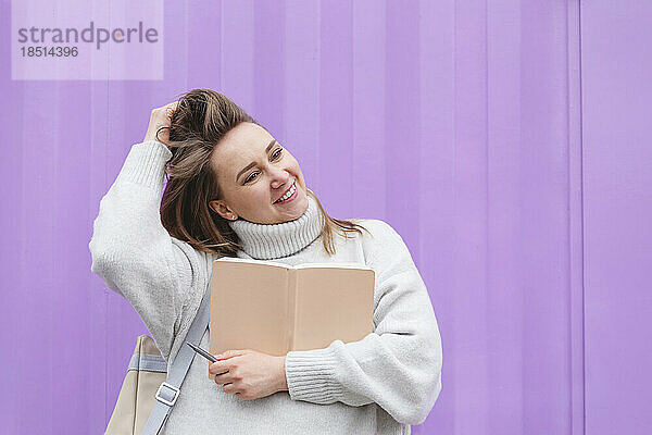 Smiling woman holding diary in front of purple wall