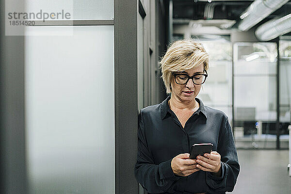 Blonde Geschäftsfrau benutzt Mobiltelefon und lehnt an der Wand im Büro