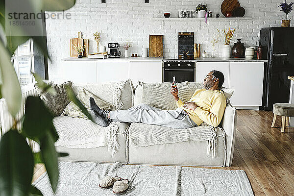 Reifer Mann benutzt Smartphone und liegt zu Hause auf dem Sofa