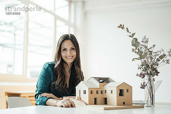 Glückliche Geschäftsfrau mit Hausmodell sitzt im Heimbüro