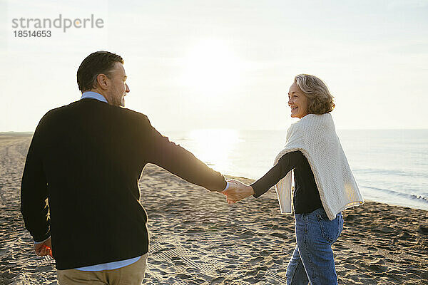 Lächelnde reife Frau hält Händchen mit Mann und geht am Strand spazieren