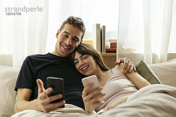Happy couple with smart phones relaxing on bed at home