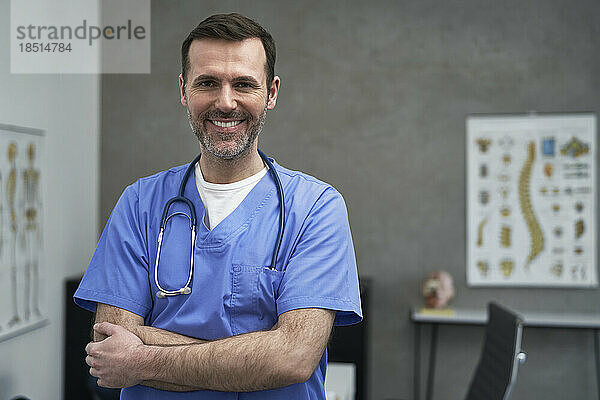 Lächelnder reifer Arzt  der mit verschränkten Armen in der Klinik steht