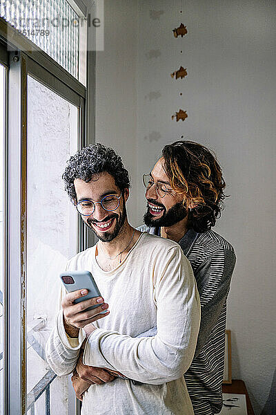 Glücklicher schwuler Mann umarmt Freund mit Smartphone am Fenster zu Hause