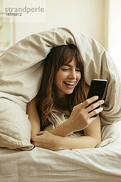 Glückliche Frau  die zu Hause ihr in eine Decke gehülltes Smartphone benutzt