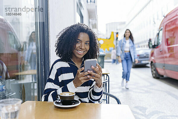 Glückliche junge Frau mit Smartphone sitzt im Straßencafé