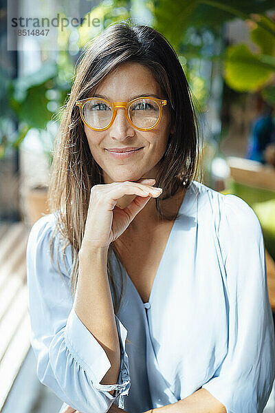 Lächelnde Frau mit Brille sitzt mit der Hand am Kinn im Café
