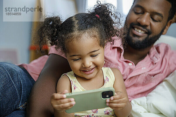 Nettes lächelndes Mädchen  das mit seinem Vater zu Hause auf das Smartphone schaut