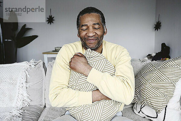 Lächelnder reifer Mann umarmt Kissen  der zu Hause auf dem Sofa sitzt
