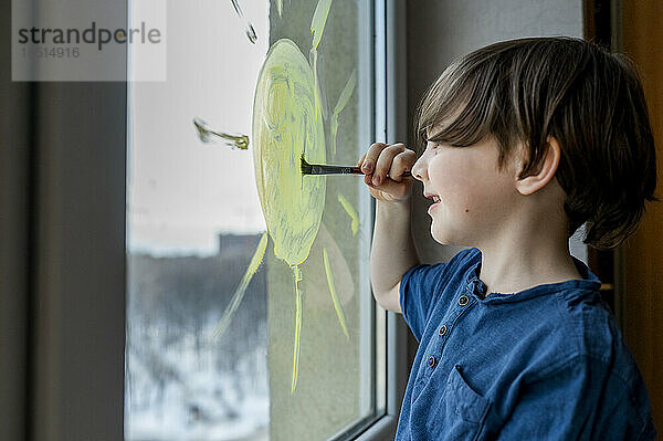 Lächelnder Junge zeichnet mit Pinsel Sonne auf Glas